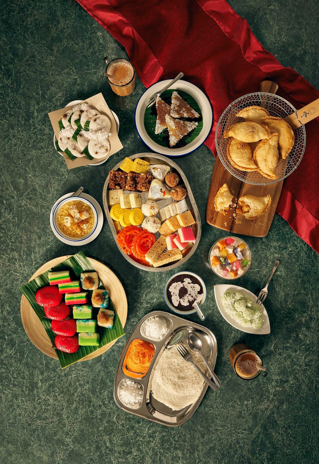 This overhead shot showcases a variety of Southeast Asian desserts and drinks arranged on a green surface with a red cloth. The spread includes colourful sweets on bamboo plates, a silver tray with various treats, and small bowls containing different desserts. Drinks are served in glasses, and the overall presentation is vibrant and appetising.